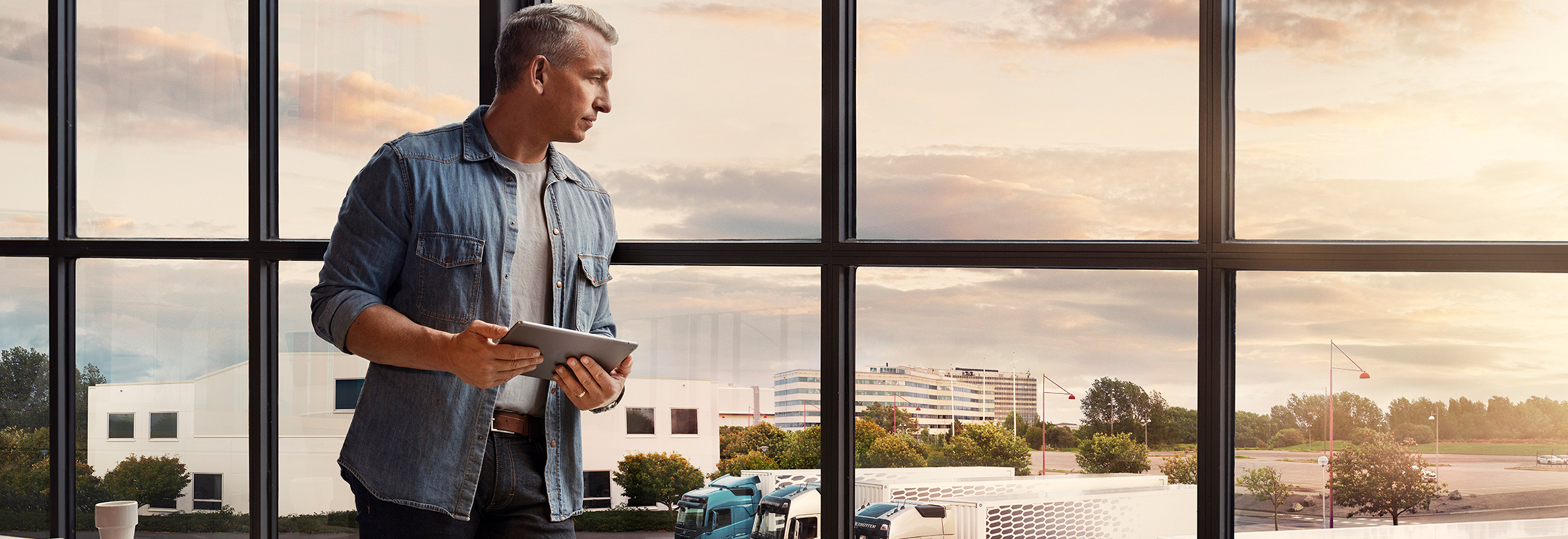 Egy táblagépet tartó férfi áll az ablaknál, miközben lenéz a saját teherautó-flottájára